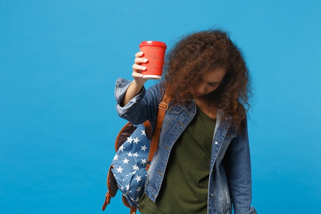 Giovane studentessa teenager della ragazza afroamericana in vestiti del denim, tazza di carta della tenuta dello zaino isolata sulla parete blu