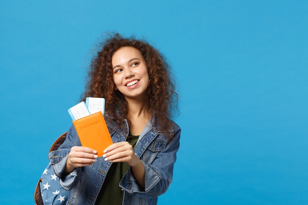 Giovane studentessa teenager della ragazza afroamericana in vestiti del denim, passaggio della tenuta dello zaino isolato sulla parete blu