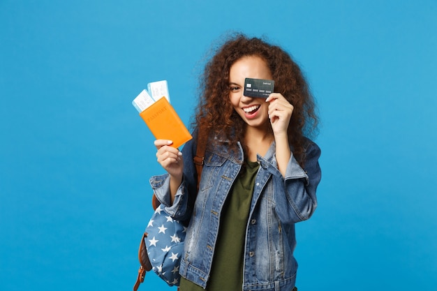 Giovane studentessa teenager della ragazza afroamericana in vestiti del denim, passaggio della tenuta dello zaino isolato sulla parete blu