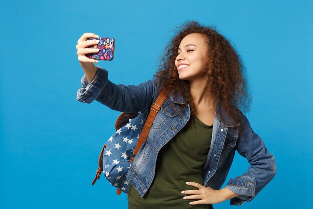 Giovane studentessa teenager della ragazza afroamericana in vestiti del denim, telefono della tenuta dello zaino isolato sulla parete blu