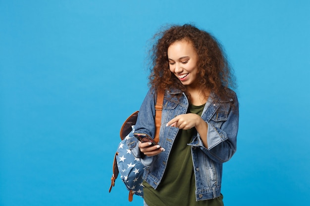 Giovane studentessa teenager della ragazza afroamericana in vestiti del denim, telefono della tenuta dello zaino isolato sulla parete blu