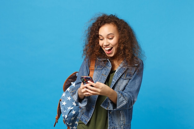 Young african american girl teen student in denim clothes, backpack hold phone isolated on blue wall