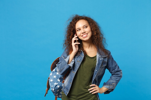 Giovane studentessa teenager della ragazza afroamericana in vestiti del denim, telefono della tenuta dello zaino isolato sulla parete blu