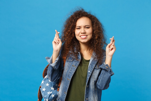 Young african american girl teen student in denim clothes, backpack making wish isolated on blue wall