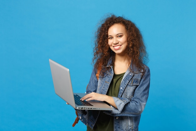 Foto giovane studentessa teenager della ragazza afroamericana in vestiti del denim, lavoro dello zaino sul pc isolato sulla parete blu