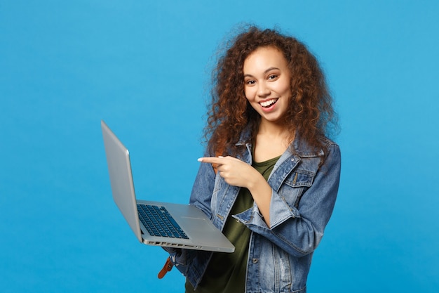 Young african american girl teen student in denim clothes, backpack work on pc isolated on blue wall