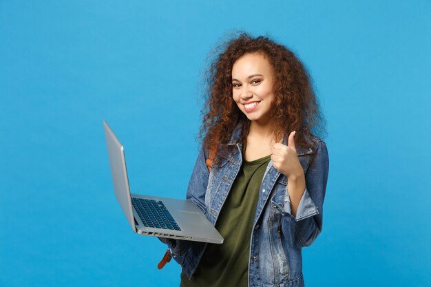 Giovane studentessa teenager della ragazza afroamericana in vestiti del denim, lavoro dello zaino sul pc isolato sulla parete blu