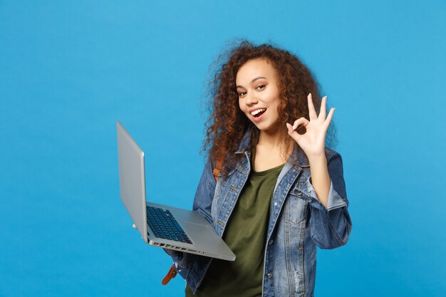 Giovane studentessa teenager della ragazza afroamericana in vestiti del denim, lavoro dello zaino sul pc isolato sulla parete blu