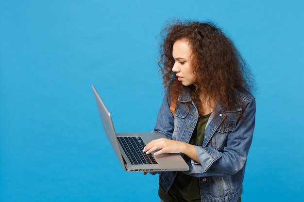 Young african american girl teen student in denim clothes, backpack work on pc isolated on blue wall