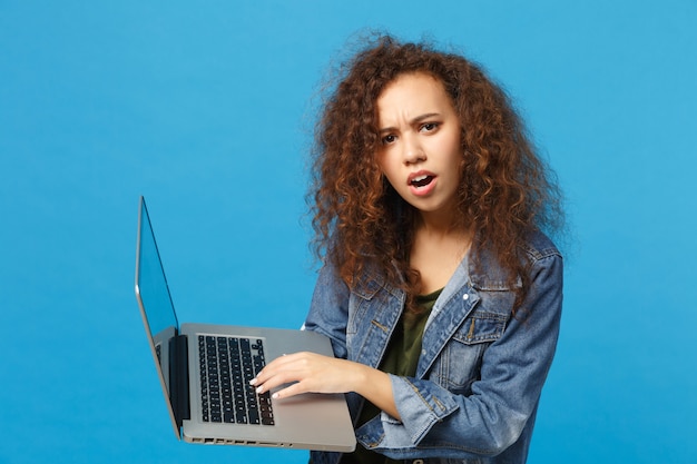 Young african american girl teen student in denim clothes, backpack work on pc isolated on blue wall