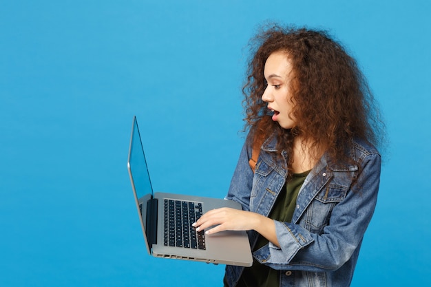 Young african american girl teen student in denim clothes, backpack work on pc isolated on blue wall