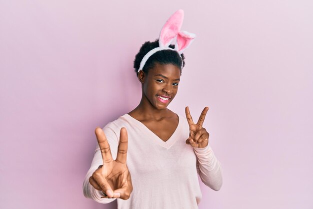 Young african american girl wearing cute easter bunny ears smiling looking to the camera showing fingers doing victory sign number two