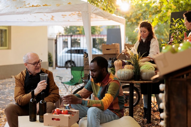 Photo young african american guy customer tasting homemade natural wine while sitting at table with winemaker at farmers market. consumer sampling local produce while visiting organic food festival