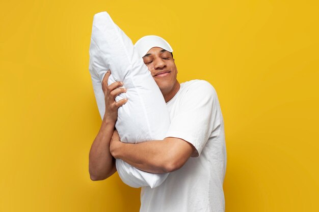 Young african american guy in pajamas and sleep mask hugs soft comfortable pillow and smiles