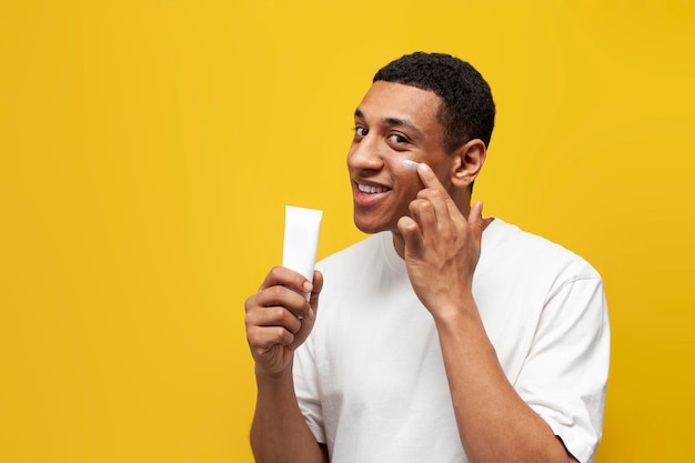 Young african american guy in white tshirt shows tube of cream on yellow isolated background