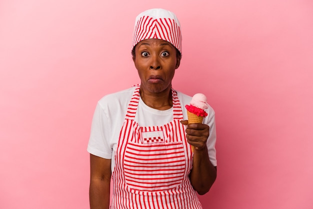Молодая афро-американская женщина-производитель мороженого, держащая мороженое на розовом фоне, пожимает плечами и смущается открытыми глазами.