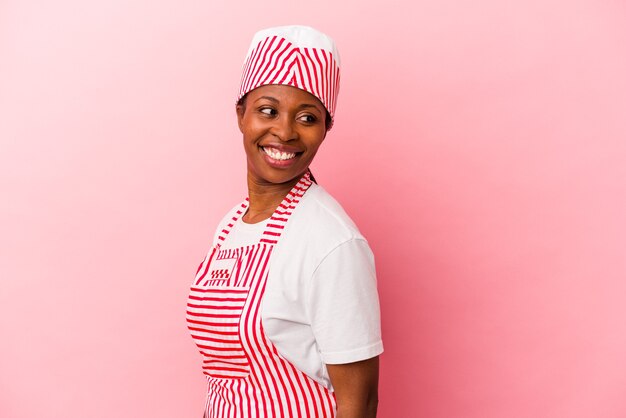 분홍색 배경에 고립된 젊은 아프리카계 미국인 아이스크림 메이커 여성은 옆으로 웃고, 명랑하고 유쾌해 보입니다.