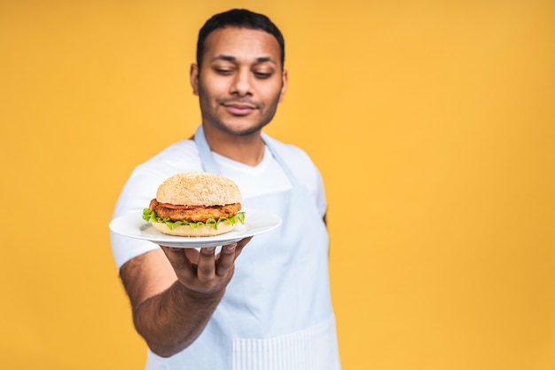 黄色の背景で隔離のハンバーガーを食べる若いアフリカ系アメリカ人のインドの黒人男性。ハンバーガーを準備して調理します。