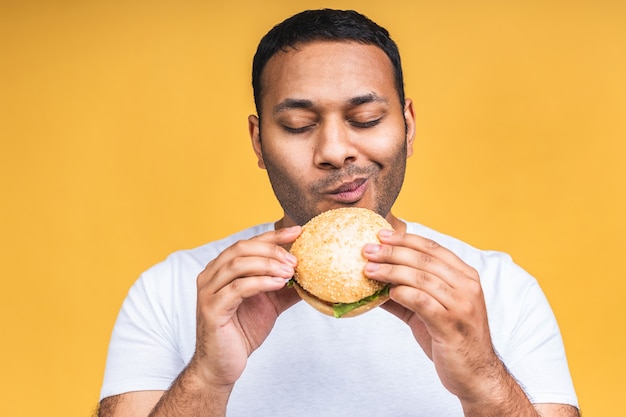 黄色の背景で隔離のハンバーガーを食べる若いアフリカ系アメリカ人のインドの黒人男性。ダイエットのコンセプト。