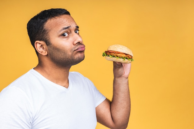 Young african american indian black man eating hamburger isolated over yellow background. Diet concept.