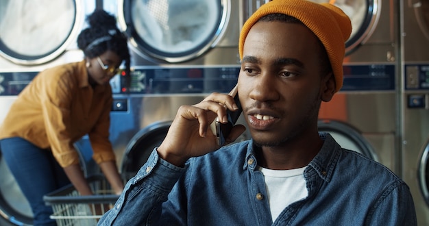 Young African American joyful handsome guy in yellow hat talking on mobile phone and smiling in laundry service room. Happy man speaking on cellphone in washhouse. Smartphone conversation.