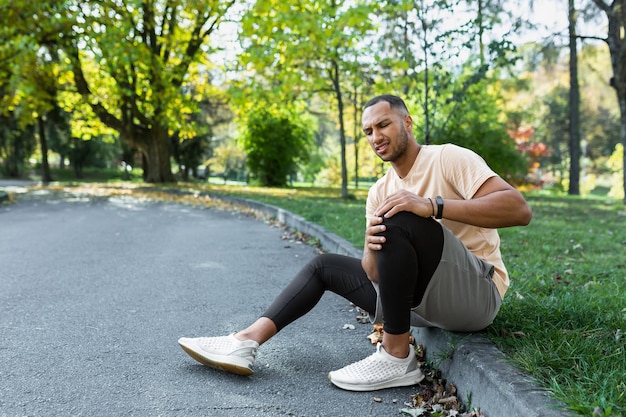 Young african american male athlete sitting in the park on a treadmill holding his knee massaging