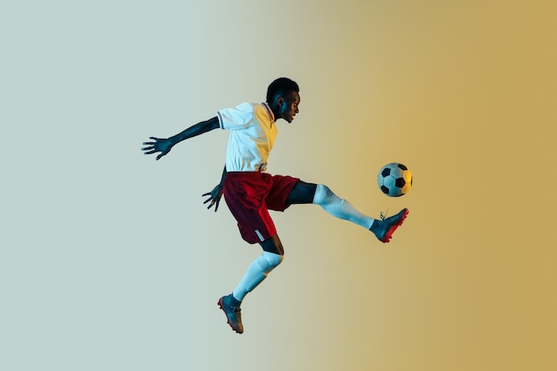 Young african-american male football or soccer player in sportwear and boots kicking ball for the goal in jump in neon light on gradient background. Concept of healthy lifestyle, professional sport.