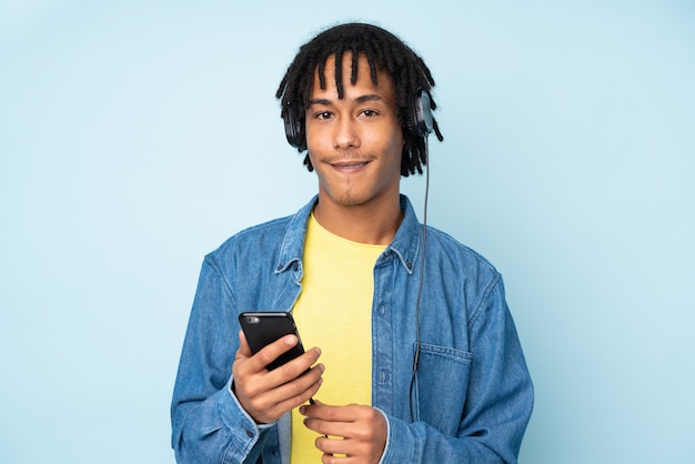 Young african american man on blue wall listening music with a mobile and looking front