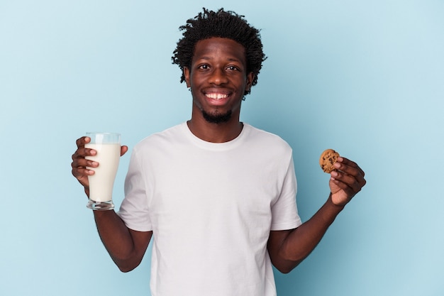 Young african american man eating chocolate chips cookies and drinking milk on blue