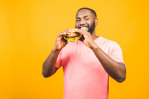 Young african american man eating hamburger isolated over yellow wall.