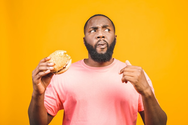 Young african american man eating hamburger isolated over yellow wall.