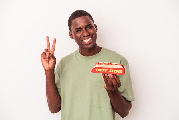 Young African American man eating a hot dog isolated on white background joyful and carefree showing a peace symbol with fingers.