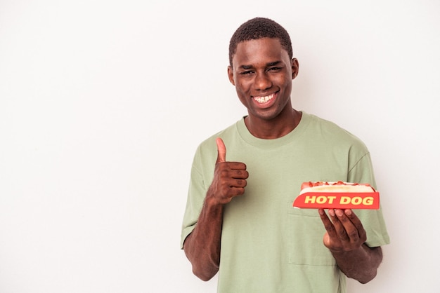 Young African American man eating a hot dog isolated on white background smiling and raising thumb up