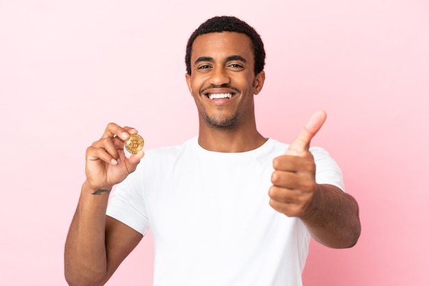 何か良いことが起こったので、親指を立てて孤立したピンクの背景の上にビットコインを保持している若いアフリカ系アメリカ人の男