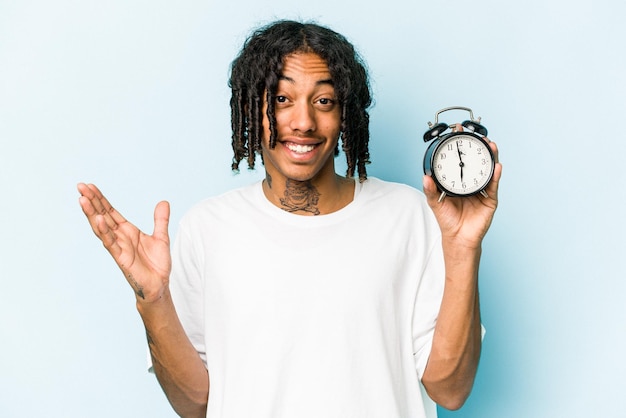 Young African american man holding alarm clock isolated on blue background receiving a pleasant surprise excited and raising hands