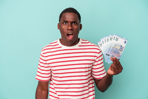 Young african american man holding a bank notes isolated on blue background screaming very angry and aggressive.