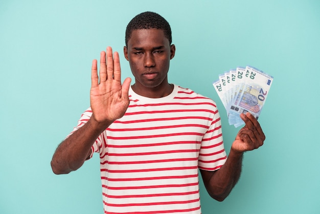 Young African American man holding a bank notes isolated on blue background standing with outstretched hand showing stop sign, preventing you.