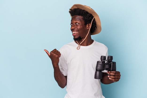 Young african american man holding binoculars isolated on blue background points with thumb finger away, laughing and carefree.