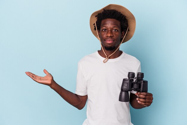 Young african american man holding binoculars isolated on blue background showing a copy space on a palm and holding another hand on waist.