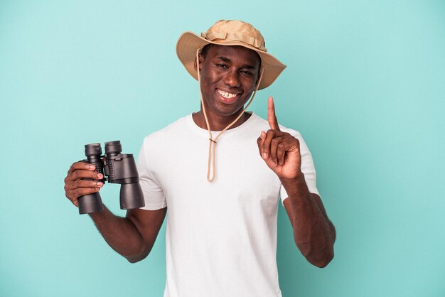 Young African American man holding binoculars isolated on blue background showing number one with finger