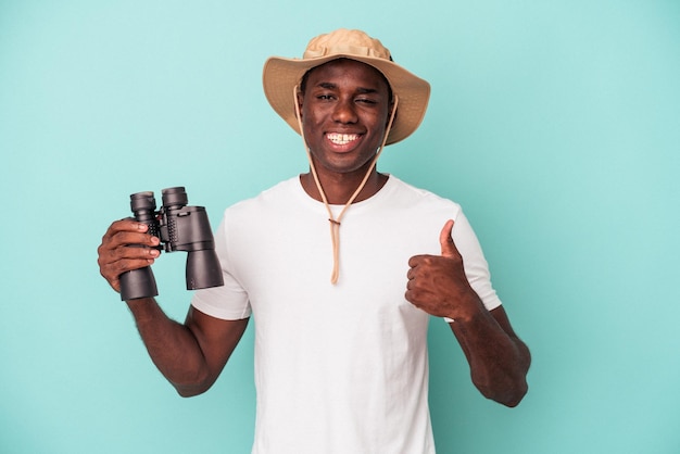 Young african american man holding binoculars isolated on blue background smiling and raising thumb up