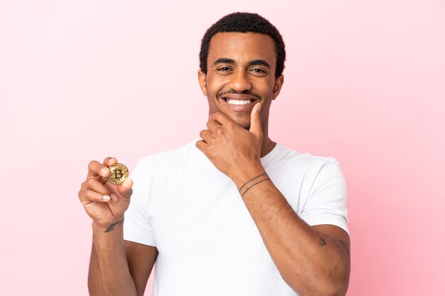 Young African American man holding a Bitcoin over isolated pink background happy and smiling