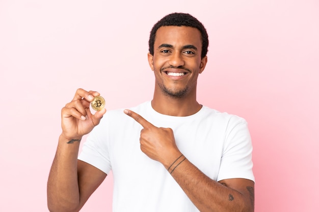 Young African American man holding a Bitcoin over isolated pink background pointing to the side to present a product