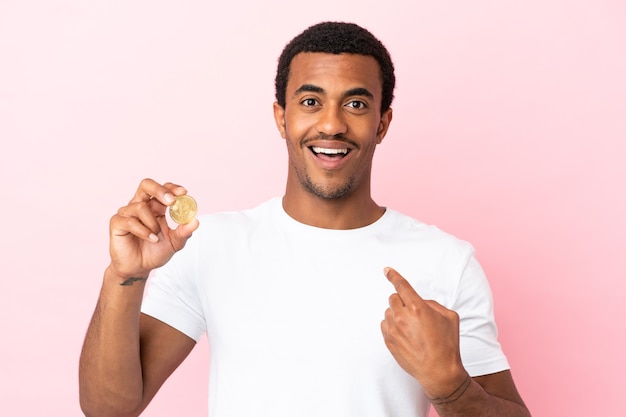 놀란 표정으로 고립된 분홍색 배경 위에 Bitcoin을 들고 있는 젊은 아프리카계 미국인 남자