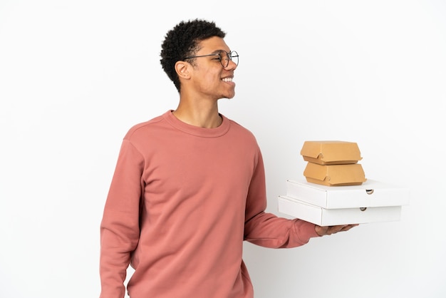 Foto giovane uomo afroamericano che tiene un hamburger e pizze isolati su fondo bianco che guarda al lato