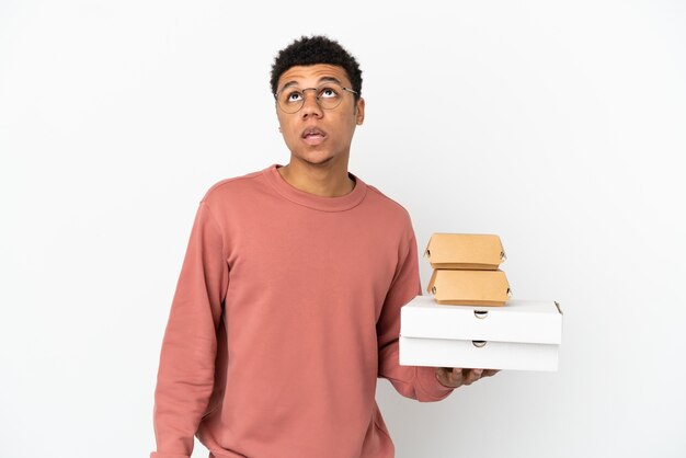 Young African American man holding a burger and pizzas isolated on white background looking up and with surprised expression