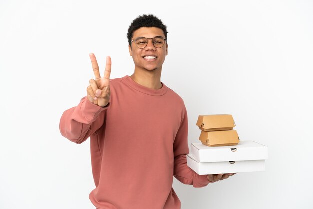 Young African American man holding a burger and pizzas isolated on white background smiling and showing victory sign