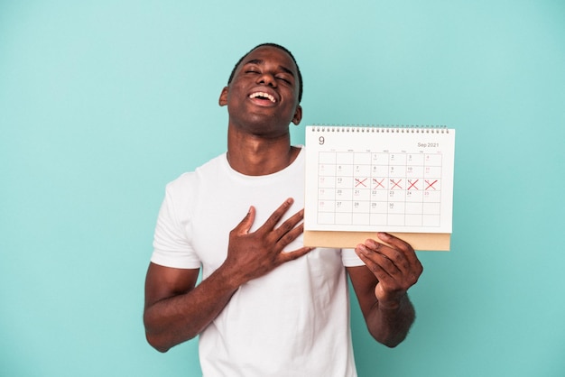 Молодой афроамериканец, держащий календарь на синем фоне, громко смеется, держа руку на груди.