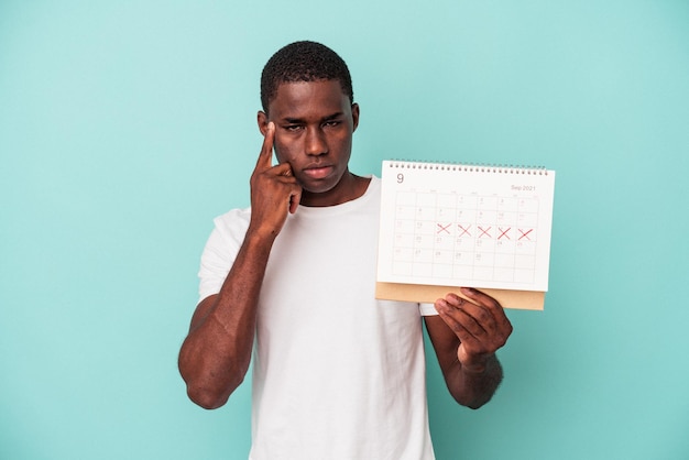 Young African American man holding a calendar isolated on blue background pointing temple with finger, thinking, focused on a task.