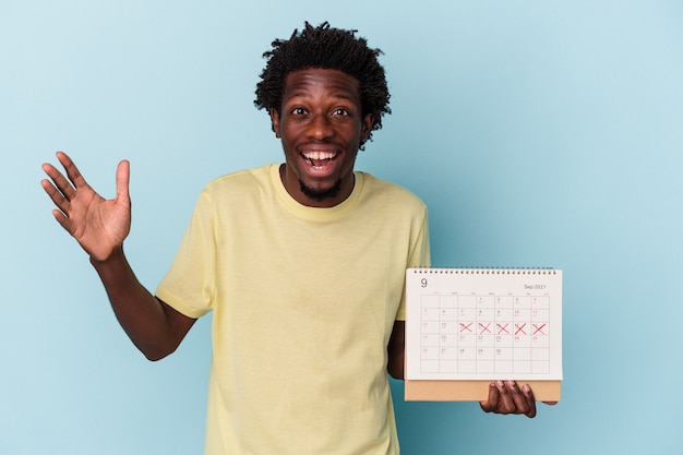 Young african american man holding calendar isolated on blue background receiving a pleasant surprise, excited and raising hands.